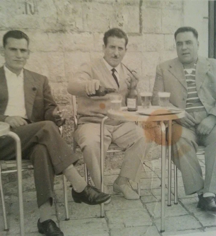 Three men in jackets sitting at an outdoor table in front of a stone wall; in the middle my grandfather Sebastiano pours from a bottle.